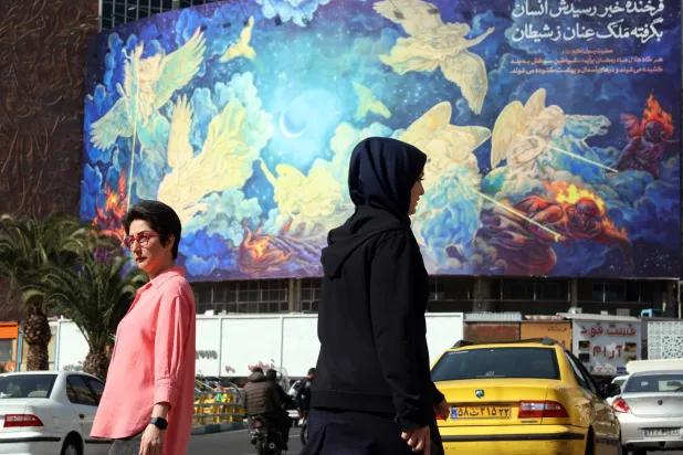 إيرانيات يسرن في طهران وخلفهن لوحة مكتوب عليها «الإنسان سوف يغلب الشيطان» (إ.ب.أ)