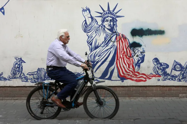 إيراني مسن يركب دراجة هوائية بجوار جدارية مناهضة للولايات المتحدة في طهران (إ.ب.أ)