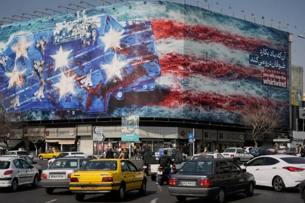 Vehicles pass a billboard depicting a US aircraft carrier with damaged fighter jets on its deck and a sign in Farsi and English reading, "If you sow the wind, you'll reap the whirlwind," at Enqelab-e-Eslami Square in Tehran, Iran, Sunday, Feb. 22, 2026. (AP) 