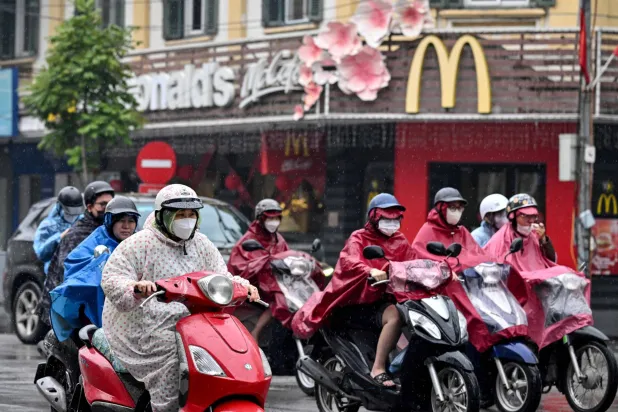People wearing raincoats ride motorbikes through the rain past a fast-food restaurant in Hanoi on February 26, 2026. (Photo by Nhac NGUYEN / AFP)