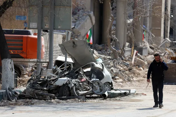رجل إيراني يسير بجوار الأنقاض والسيارات المحطمة بوسط طهران (إ.ب.أ)