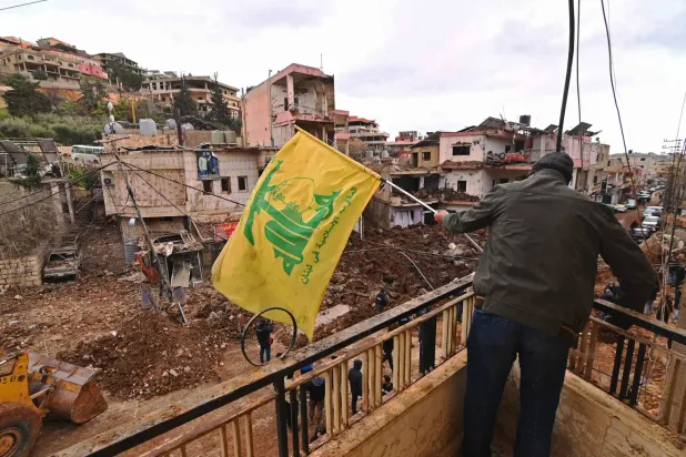 This photograph taken during a media tour organized by the Hezbollah shows a man installing a flag of Hezbollah on the balcony of a damaged building at Nabi Sheet town after an Israeli military operation in the Bekaa Valley of Lebanon, on March 7, 2026.  (Photo by FADEL itani / AFP)