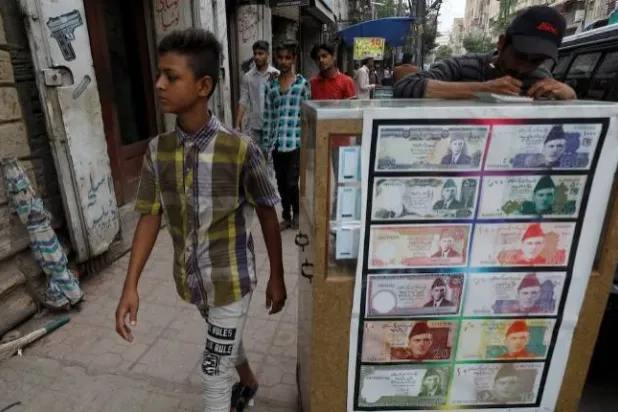فتى يمر بجوار كشك صرافة مزين بصور الأوراق النقدية في كراتشي (رويترز)