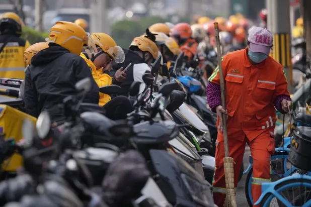  A street cleaner walks by food delivery riders gather outside restaurants waiting for their online orders, in Beijing, China, Wednesday, March 25, 2026. (AP) 