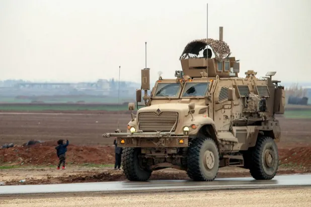 In this file photo dated Feb 7, 2026, boys along a road wave to an approaching US military mine-resistant ambush protected (MRAP) armored fighting vehicle moving in a convoy transporting ISIS group detainees being transferred to Iraq from Syria, on the outskirts of Qahtaniyah in Syria's northeastern Hasakeh province. (AFP) 