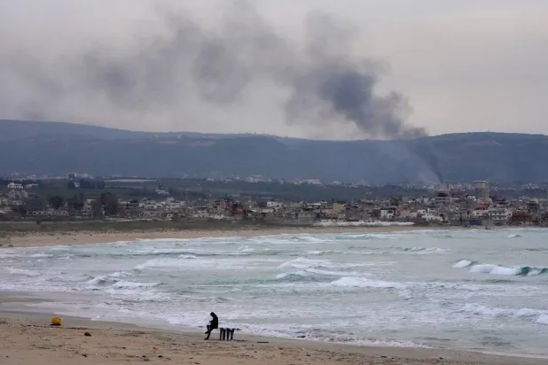 A man checks his phone on a beach as smoke rises from Israeli artillery shells on Qlaileh village, visible from Tyre city, south Lebanon, March 28, 2026. (AP)