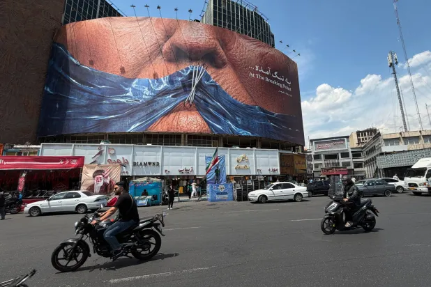 سيارات تمر قرب لوحة دعائية في وسط طهران تُظهر رسماً لمضيق هرمز وشفتي الرئيس الأميركي دونالد ترمب مخيطتين (أ.ب)