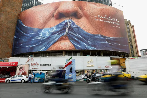 إيرانيون يمرون أمام لوحة دعائية مناهضة للولايات المتحدة تُظهر رسماً لمضيق هرمز وشفتي الرئيس الأميركي دونالد ترمب مخيطتين (رويترز)