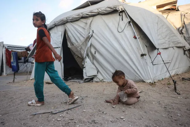 A Palestinian girl walks past a toddler playing among makeshift shelters in Khan Yunis in the southern Gaza Strip on May 21, 2026. Since October 10, a fragile US-sponsored truce in Gaza has largely halted the fighting between Israeli forces and Hamas, but both sides have alleged frequent violations. (Photo by Bashar Taleb / AFP)