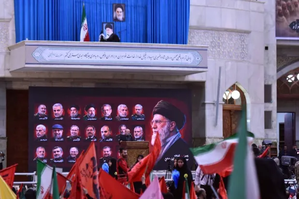 كرسي فارغ تعلوه صورة المرشد السابق علي خامنئي في موقع مراسم ذكرى وفاة الخميني جنوب طهران، وأدناها لوحة تضم صور قادة ومسؤولين إيرانيين قُتلوا خلال الحرب، بينما رُفعت أعلام الجمهورية الإسلامية ورايات حمراء بين الحشود المشاركة في المراسم (جماران)