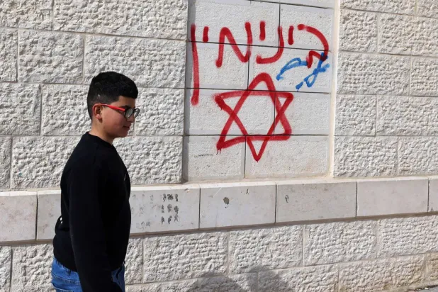 Pazar günü Batı Şeria'nın Nablus şehrinin güneyindeki Cellud köyünde yerleşimci saldırısında hasar gören bir evin duvarlarına yazılmış hakaret içerikli İbranice yazıların önünden geçen Filistinli bir çocuk (AFP)