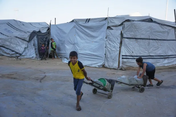 أطفال فلسطينيون نازحون ينقلون حاويات المياه في ملعب اليرموك لكرة القدم الذي تضرر خلال الحرب الإسرائيلية بمدينة غزة (أ.ب)