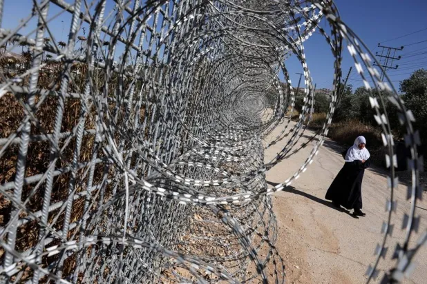  Jawaher Foqahaa walks past the tall metal fencing installed by the Israeli military that closed off four of the five entrances to their town and runs directly past the family home, in Sinjil, near Ramallah, in the Israeli-occupied West Bank, June 26, 2026. (Reuters)