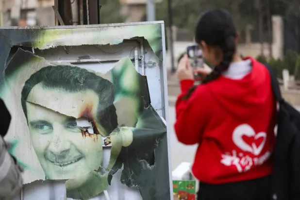 A woman uses her mobile phone near a damaged picture of ousted Syrian ruler Bashar al-Assad as people celebrate, after the Syrian opposition announced that they have ousted President Bashar al-Assad, in Qamishli, Syria December 8, 2024. REUTERS/Orhan Qereman