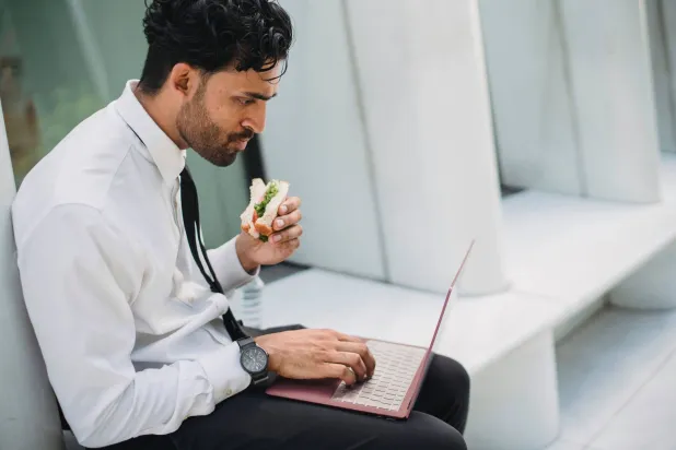 شخص يتناول الطعام أثناء استخدامه للكمبيوتر المحمول (بيكسلز)
