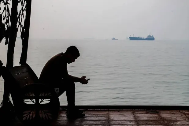 A man checks his cellphone at a waterfront shelter as commercial vessels appear anchored in the Strait of Hormuz off Bandar Abbas, Iran, Monday, Tuesday, Oct, 6, 202 (AP)