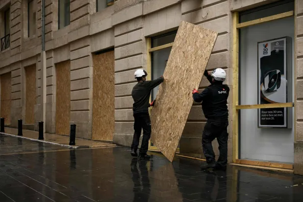 Workers in hard hats lift a wooden panel to board up a shop window ahead of a demonstration planned for October 24, in central Bordeaux, southwestern France on October 22, 2026. (AFP)