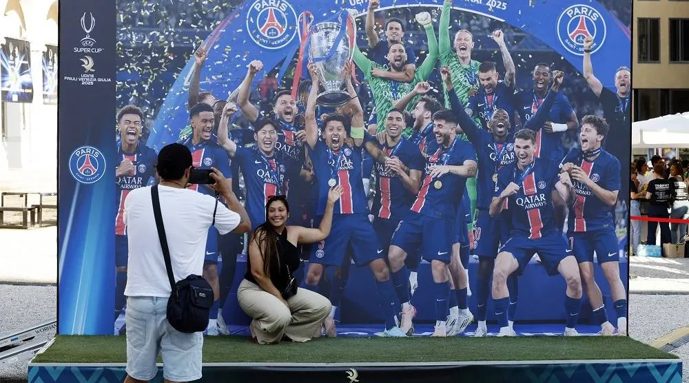 Football - UEFA Super Cup - Final - Paris St Germain v Tottenham Hotspur - Preview - Udine, Italy - August 12, 2025. People pose for photos in front of an image of Paris St Germain lifting the trophy after they won the Champions League. (Reuters) 