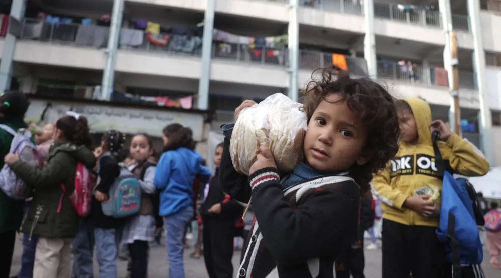 طفلة فلسطينية تحمل خبزاً في حين يستعد آخرون لدخول صفوفهم في مدرسة للأونروا تؤوي عائلات نازحة تضطر يومياً لإخراج أمتعتها لمتابعة يوم دراسي جديد في دير البلح وسط قطاع غزة السبت (أ.ف.ب)