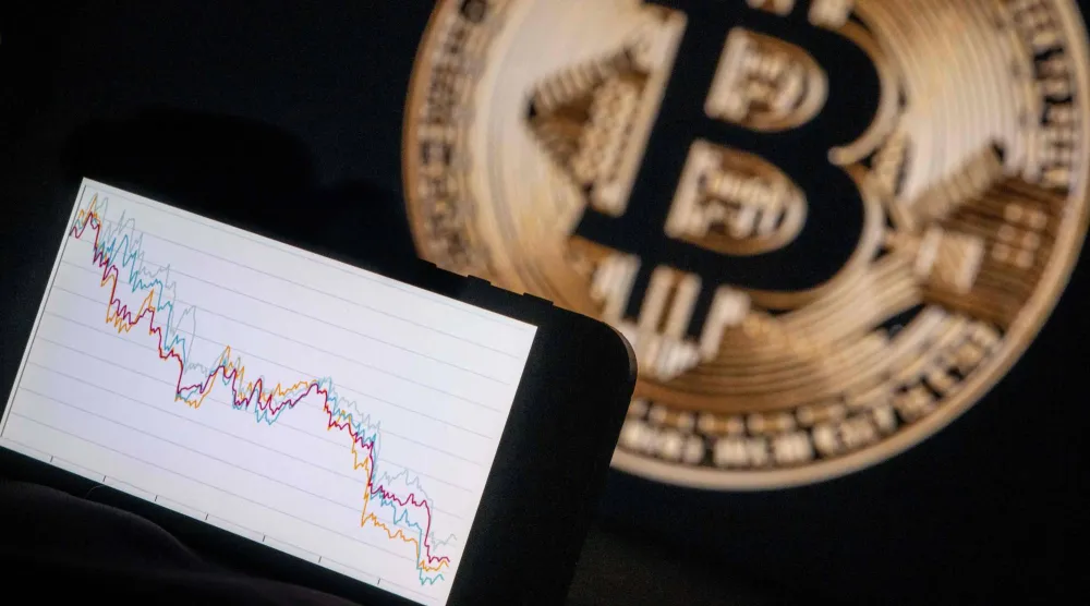 تُظهر هذه الصورة التوضيحية هاتفاً يعرض اتجاهاً هابطاً في سوق الأسهم أمام شاشة تُظهر شعار «بتكوين» (أ.ف.ب)