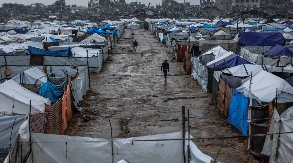 فلسطينيون يسيرون على طول زقاق موحل في مخيم مؤقت يؤوي نازحين فلسطينيين بعد هطول أمطار غزيرة بحي الزيتون بمدينة غزة (أ.ف.ب)