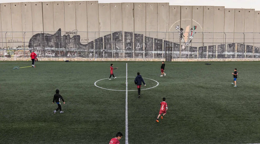 أطفال مخيم عايدة للاجئين الفلسطينيين يتدربون على كرة القدم في ملعب صغير ملاصق لجدار الفصل بالضفة الغربية المحتلة (أ.ف.ب)