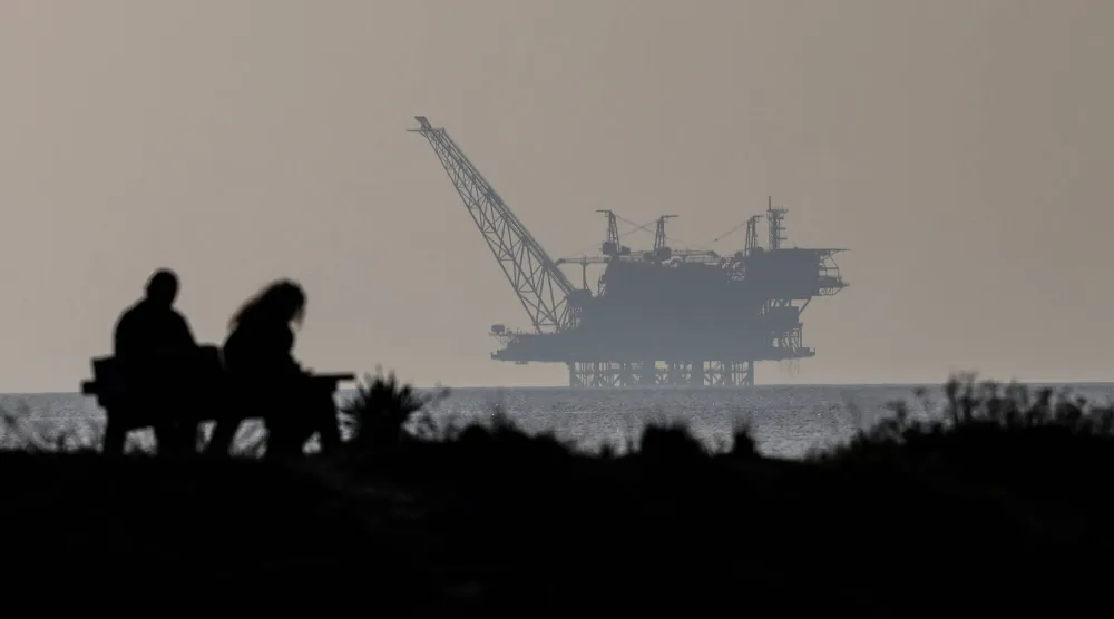 شخصان يجلسان على مقعد وخلفهم حقل غاز ليفياثان في محمية شاطئ هابونيم الطبيعية شمال إسرائيل (إ.ب.أ)