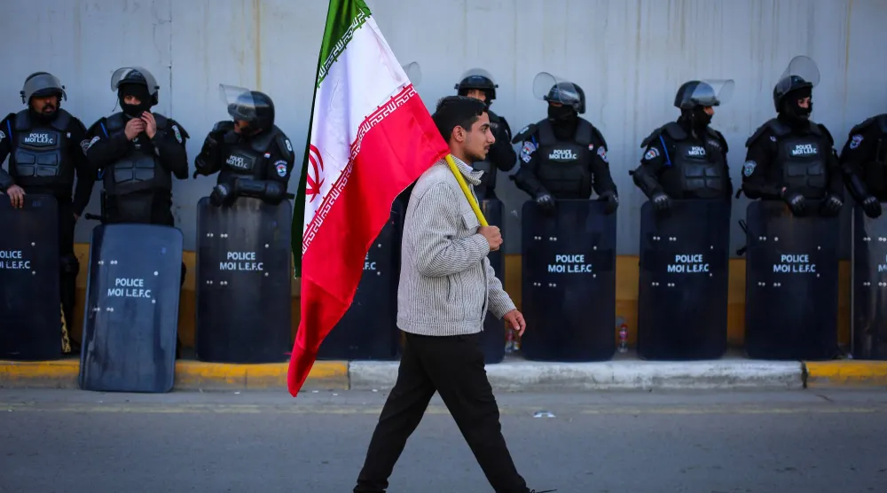 عراقي يحمل علم إيران يمرّ أمام قوات الأمن أمام السفارة الإيرانية في بغداد خلال مظاهرة لإظهار التضامن ضد التهديدات الأميركية (د.ب.أ)