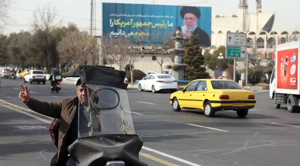 رجل إيراني يرفع علامة النصر أثناء مروره بجوار صورة للمرشد علي خامنئي في أحد شوارع طهران (إ.ب.أ)