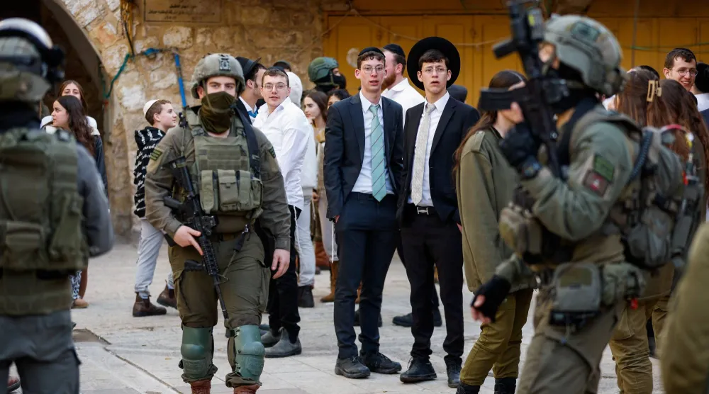 مستوطنون إسرائيليون يراقبون في حين يقف جنود إسرائيليون حراساً خلال جولة أسبوعية في الخليل بالضفة الغربية المحتلة يوم 31 يناير 2026 (رويترز)