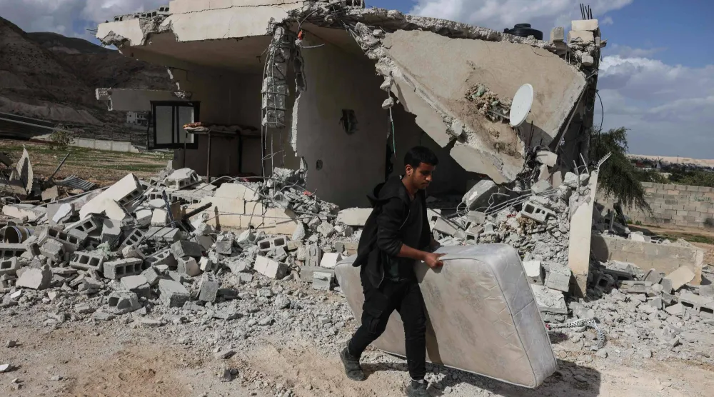 فلسطيني يحمل فراشاً ماراً بأنقاض منزل أفادت التقارير بهدمه على يد مستوطنين إسرائيليين في اليوم السابق بقرية على مشارف أريحا بالضفة الغربية المحتلة (أ.ف.ب)