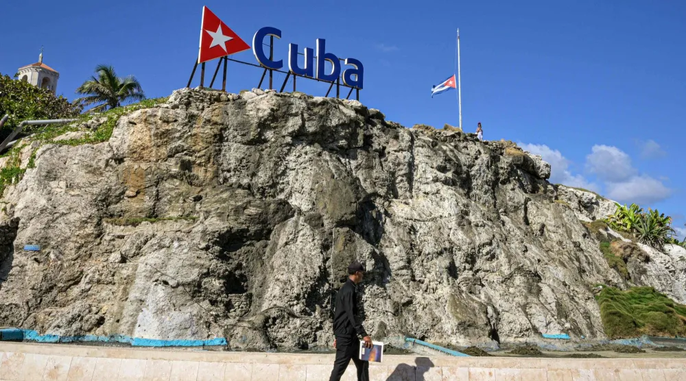 رجل كوبي يمر بجوار لافتة تحمل اسم كوبا وعَلم وطني منكس في هافانا (أ.ف.ب)