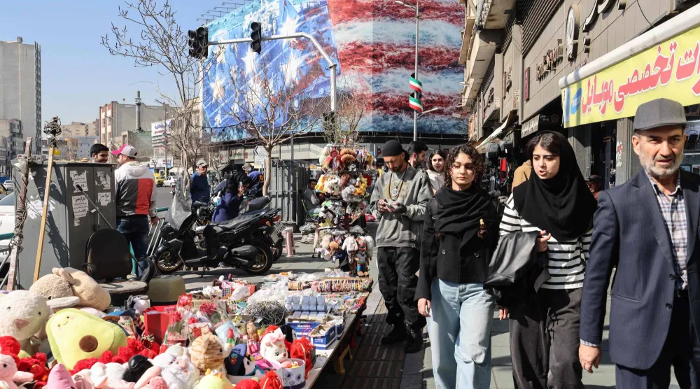 إيرانيون يمرون أمام سلع يعرضها بائع متجول مع لافتة معادية للولايات المتحدة تظهر في الخلفية بساحة انقلاب (الثورة) وسط طهران (أ.ف.ب)