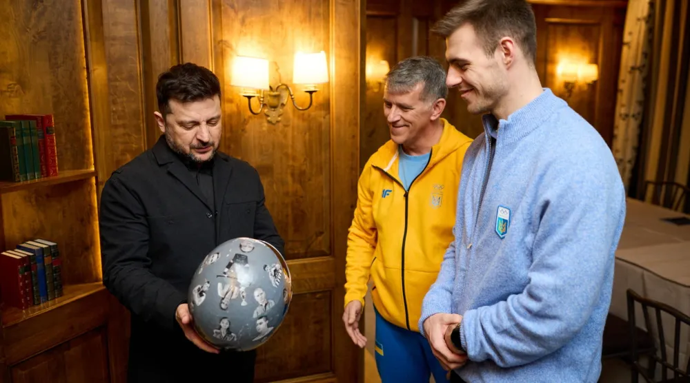 Ukrainian President Volodymyr Zelenskiy holds helmet as he meets with a Ukrainian skeleton racer Vladyslav Heraskevych , who was disqualified from the Olympic skeleton competition over his "helmet of remembrance" depicting athletes killed since Russia's invasion and his father and coach, Mykhailo Heraskevych, amid Russia's attack on Ukraine, in Munich, Germany February 13, 2026. (Ukrainian Presidential Press Service/Handout via Reuters) 