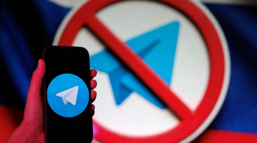 شخص يتخذ وضعية لالتقاط صورة وهو يحمل هاتفاً ذكياً بجوار شاشة تعرض شعار تطبيق المراسلة «تلغرام» على خلفية علم روسيا (رويترز)