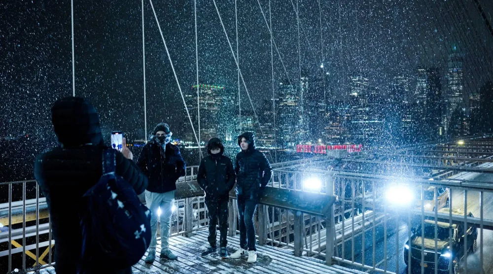 أشخاص يلتقطون الصور على جسر بروكلين أثناء تساقط الثلوج خلال عاصفة شتوية في مدينة نيويورك (رويترز)