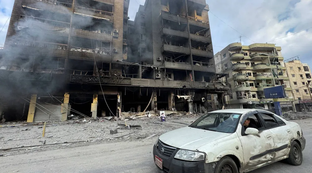 لبناني يقود سيارته بجوار مبانٍ متضررة ويتصاعد منها الدخان جراء الغارات الإسرائيلية على الضاحية الجنوبية لبيروت (رويترز)