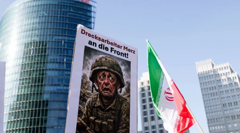 لافتة تحمل صورة المستشار الألماني فريدريش ميرتس خلال مسيرة تحت شعار «لا للحرب على إيران... السلام بدلاً من التضخم الناجم عن الحرب» بجانب العلم الإيراني في برلين (إ.ب.أ)