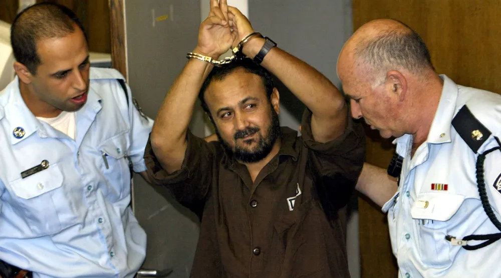 مروان البرغوثي يلوّح بيده فيما تقوم الشرطة الإسرائيلية بإحضاره إلى المحكمة لحضور جلسة النطق بالحكم في تل أبيب 20 مايو 2004 (رويترز)