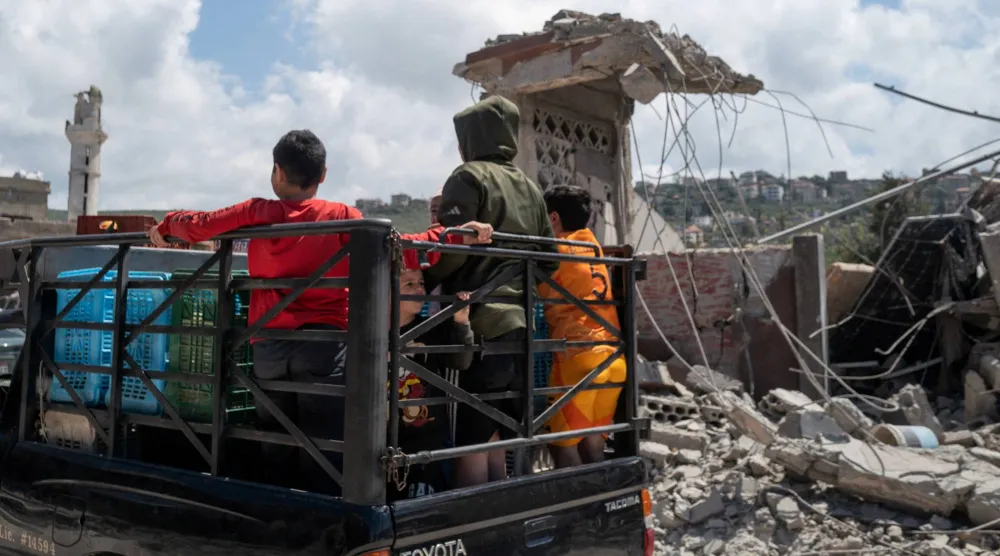 أطفال يقفون على متن شاحنة صغيرة أثناء مرورهم بجانب مبانٍ متضرّرة جرّاء غارة إسرائيلية في بلدة المنصوري جنوب لبنان (رويترز)