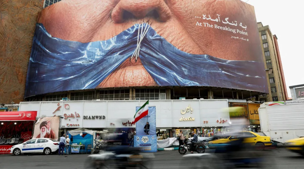 إيرانيون يمرون أمام لوحة دعائية مناهضة للولايات المتحدة تُظهر رسماً لمضيق هرمز وشفتي الرئيس الأميركي دونالد ترمب مخيطتين (رويترز)