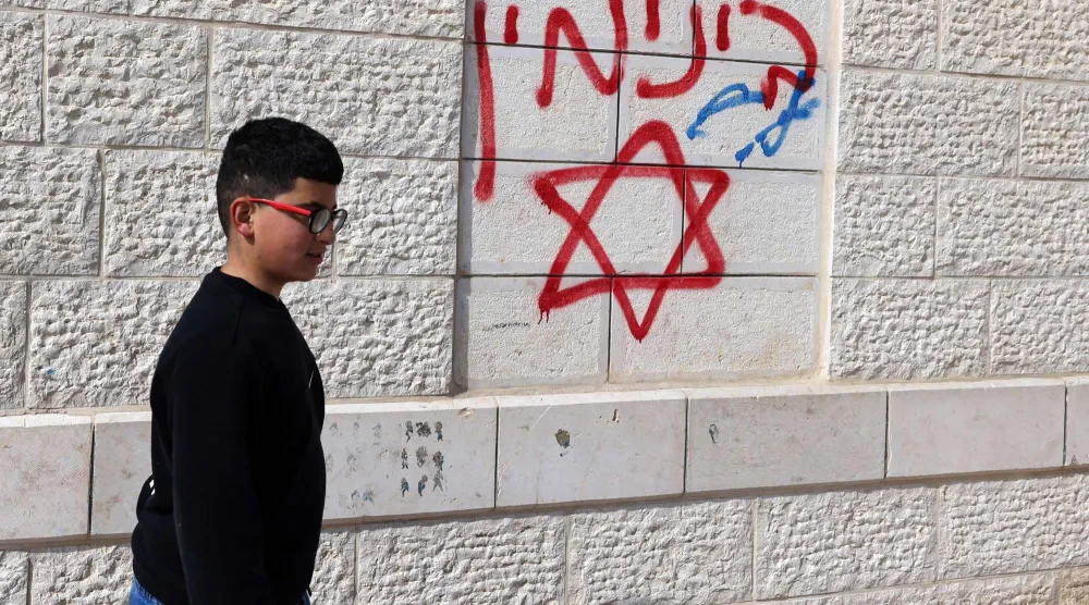 طفل فلسطيني يمرّ يوم الأحد بجوار كتابات عبرية مسيئة على جدران منزل متضرر من هجوم المستوطنين في قرية جلود جنوب نابلس بالضفة الغربية (أ.ف.ب)