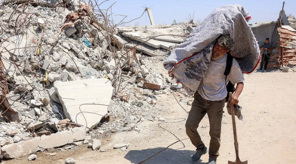 فلسطيني يحمل أمتعة على كتفه ويمر بمحاذاة الأنقاض في مخيم للنازحين بغزة (أ.ف.ب)
