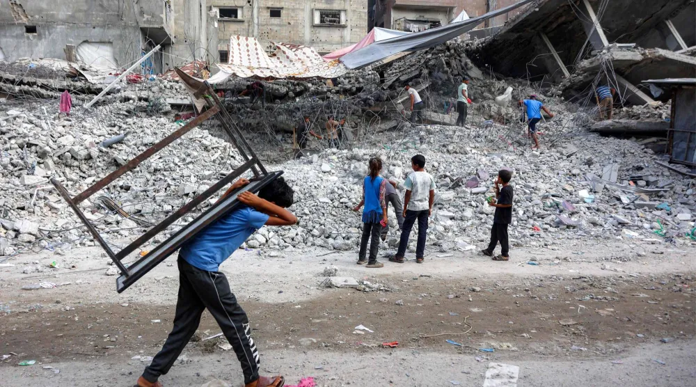 فتى فلسطيني يحمل طاولة على ظهره مارّاً بجوار عمال يقومون بتفكيك ركام مبنى دُمّر خلال الحرب الإسرائيلية ضد حركة «حماس» في غزة لاستخدام المواد المُعاد تدويرها منه مستقبلاً (أ.ف.ب)