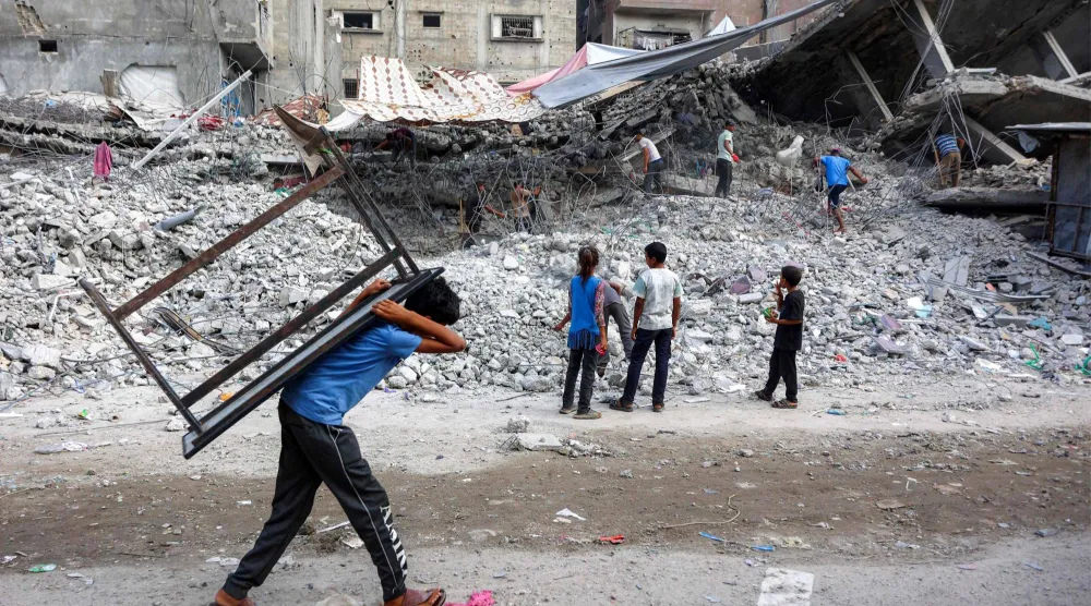 صبي فلسطيني يحمل طاولة يمر قرب عمال يجمعون أنقاض مبان دمرتها إسرائيل في الحرب على غزة (أ.ف.ب)