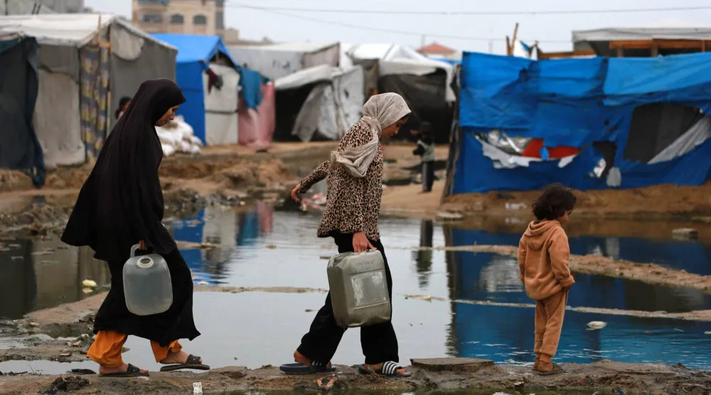 تستخدم نساء فلسطينيات نازحات طريقاً جافاً لنقل المياه إلى خيمتهن بعد هطول أمطار غزيرة شمال دير البلح في وسط قطاع غزة (أ.ف.ب)