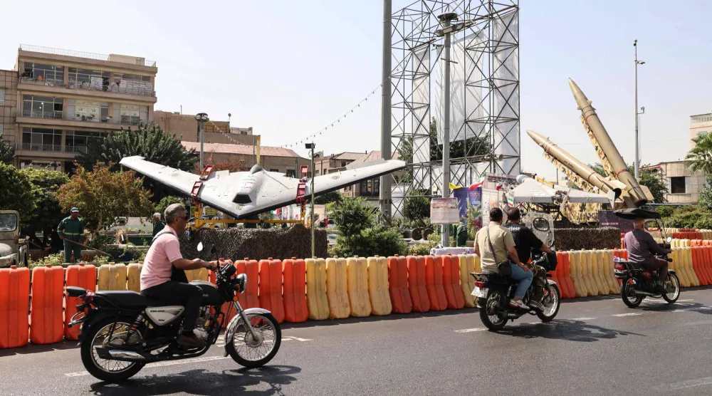 أشخاص على دراجات يمرون بجوار طائرات مُسيرة عسكرية معروضة في ساحة بهارستان بطهران (أ.ف.ب)