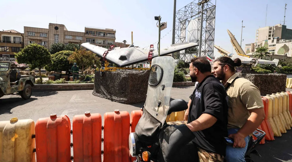 أشخاص على دراجات يمرون بجوار طائرات مُسيرة عسكرية معروضة في ساحة بهارستان بطهران (أ.ف.ب)