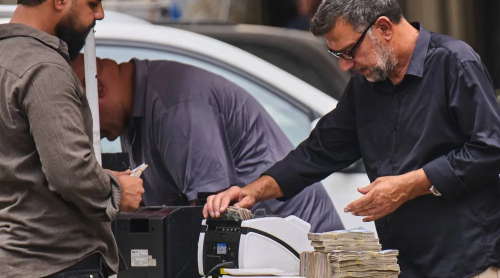 صراف عملات يقوم بعدّ رزم من أوراق الدينار العراقي خلال خدمة أحد الزبائن بمحل صرافة على جانب الطريق في بغداد (أ.ب)